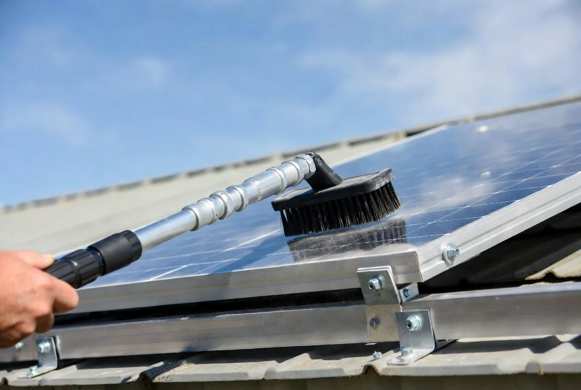 Hand using a telescoping pole with a soft nylon brush to clean dust off a rooftop solar panel on a manufactured home