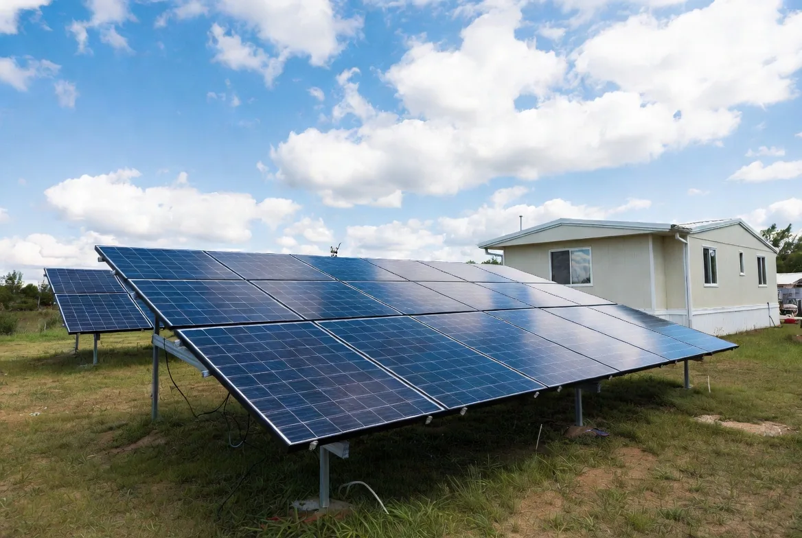 Ground-mounted solar panel array in a grassy yard next to a modern single-wide manufactured home under a clear blue sky